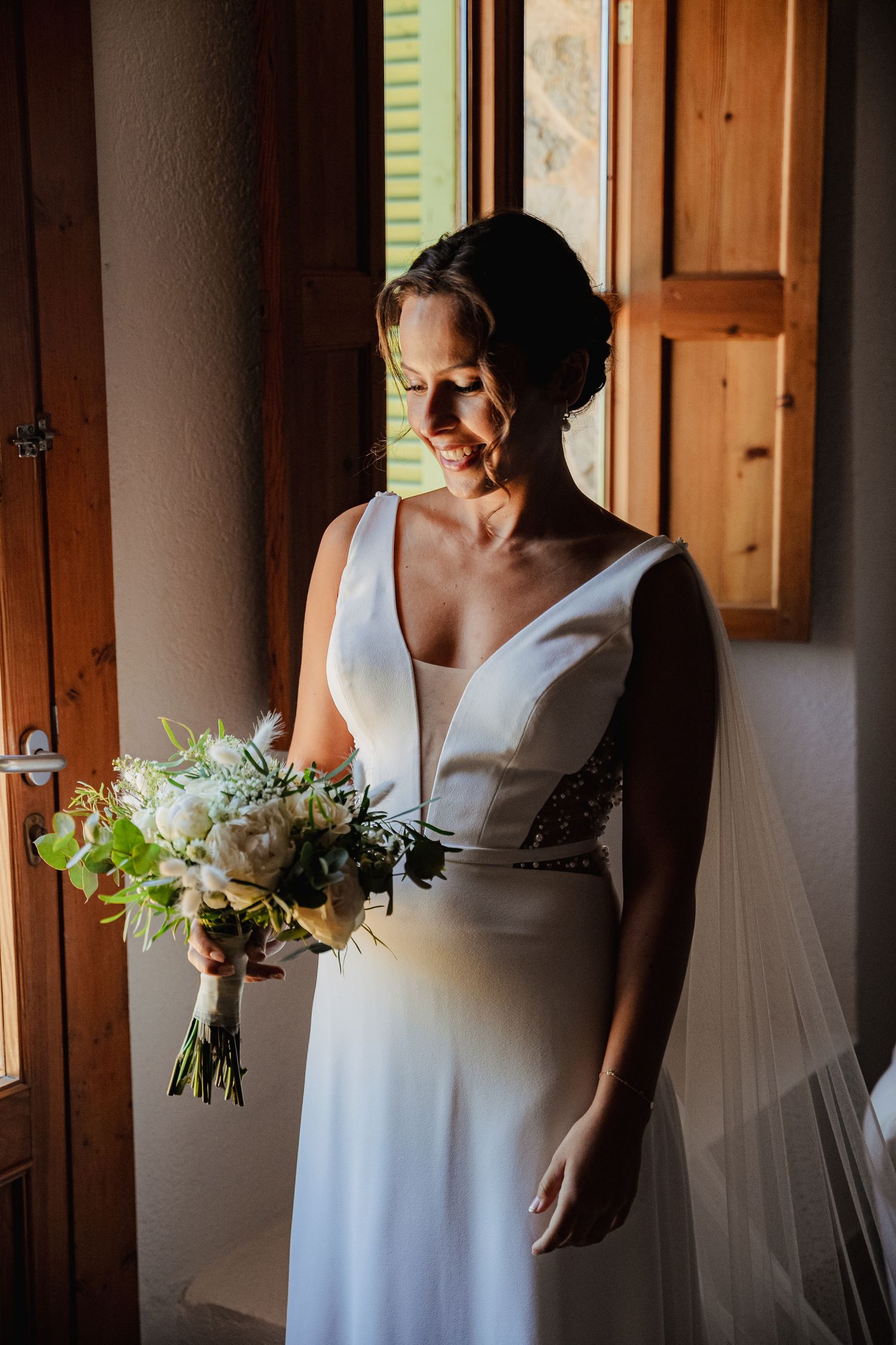 Smiling bride in deep V-neck white gown holding white floral bouquet by a rustic wooden window.