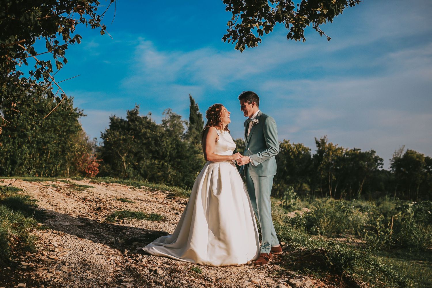Bride and groom holding hands and gazing at each other on a sunny outdoor path.