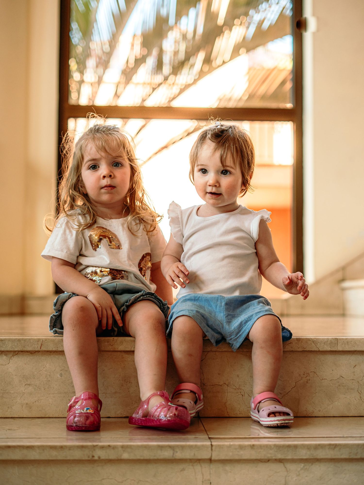 Two little girls in white tops and blue shorts sitting together on marble steps near a window.