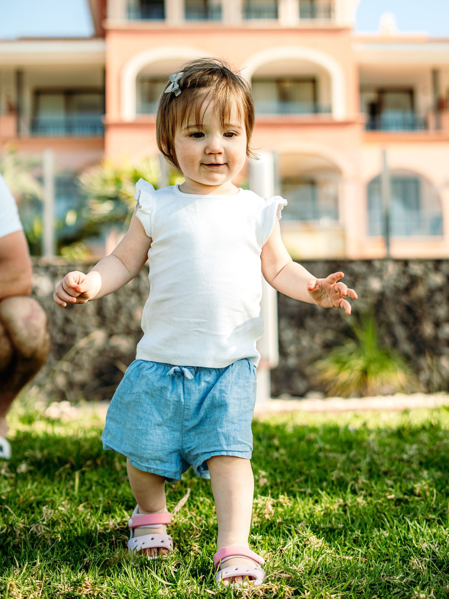 Toddler girl in white top and blue shorts walking on green grass with a building in the background.