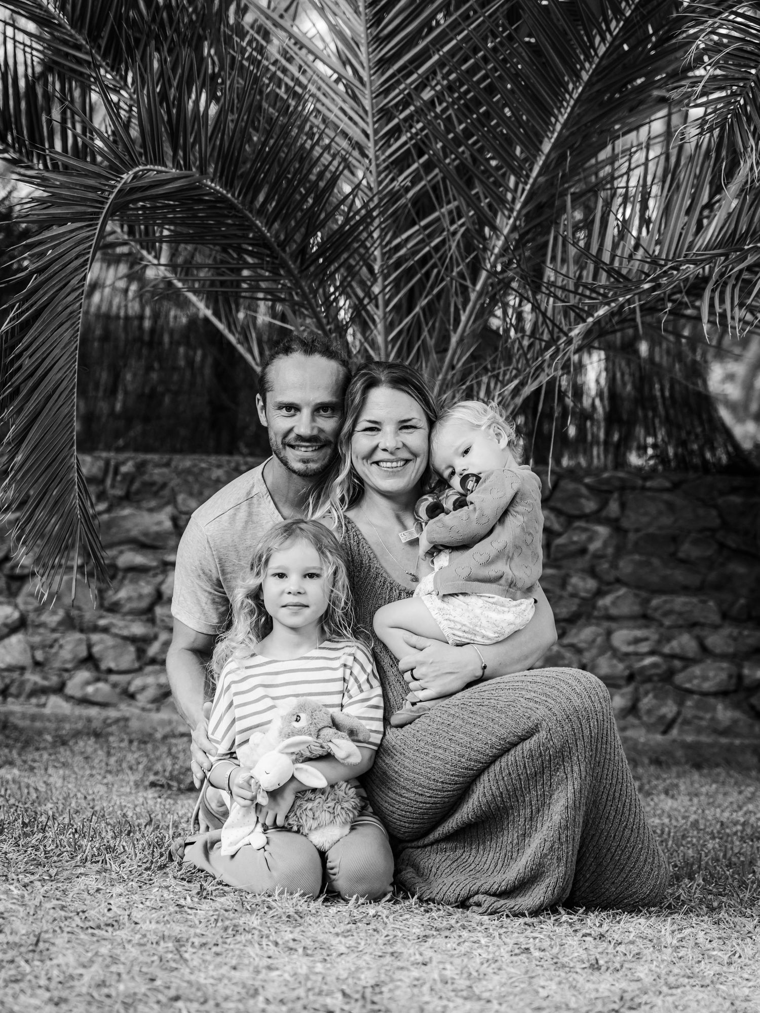 Black and white family portrait with parents and two young children under palm tree.
