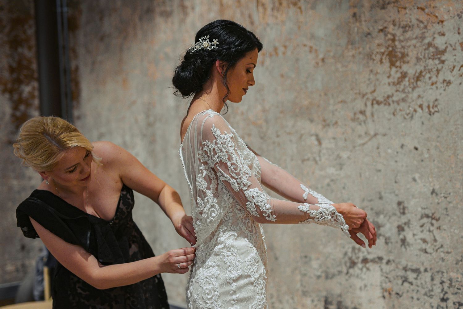 Blonde bridesmaid in black dress assists bride with lace wedding gown buttons.