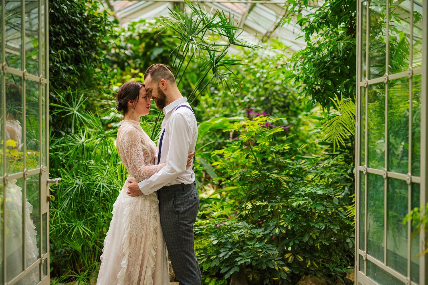 Newlyweds holding each other tenderly inside a glass greenhouse filled with vibrant tropical plants.