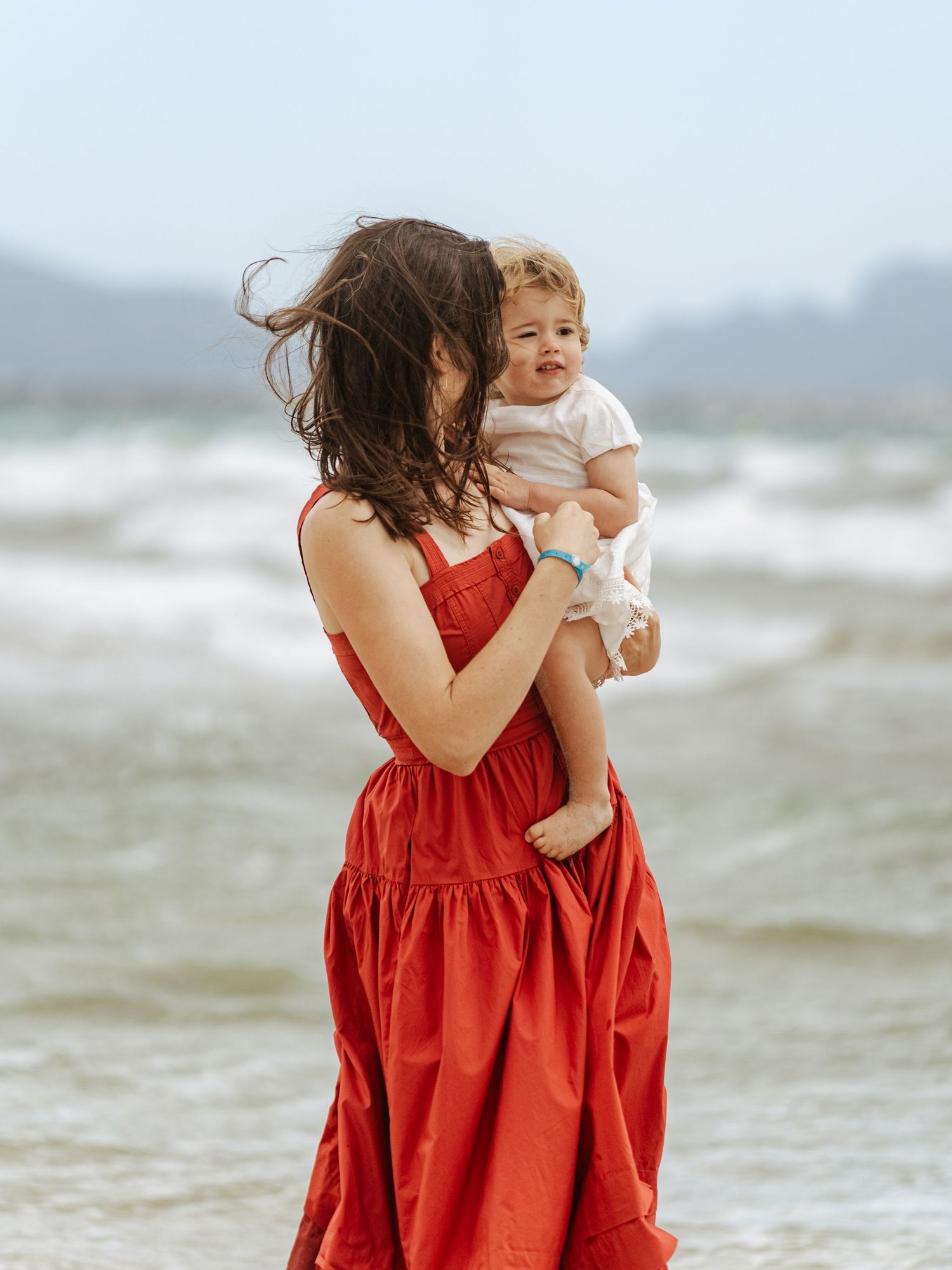 Mother in red dress holding a baby on a windy beach with ocean waves in the background.