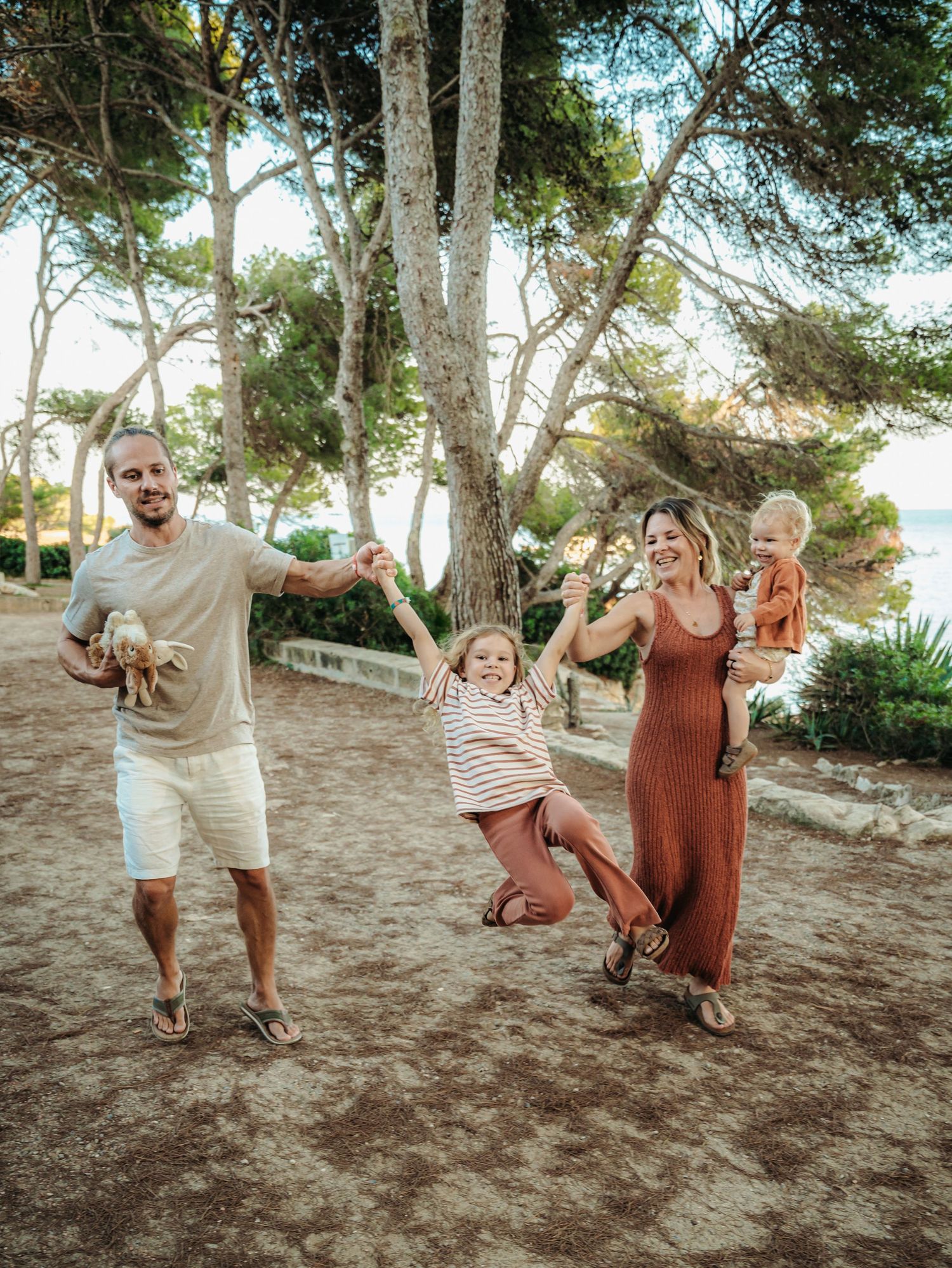 Joyful family playing outdoors under pine trees with child jumping and parents laughing together.