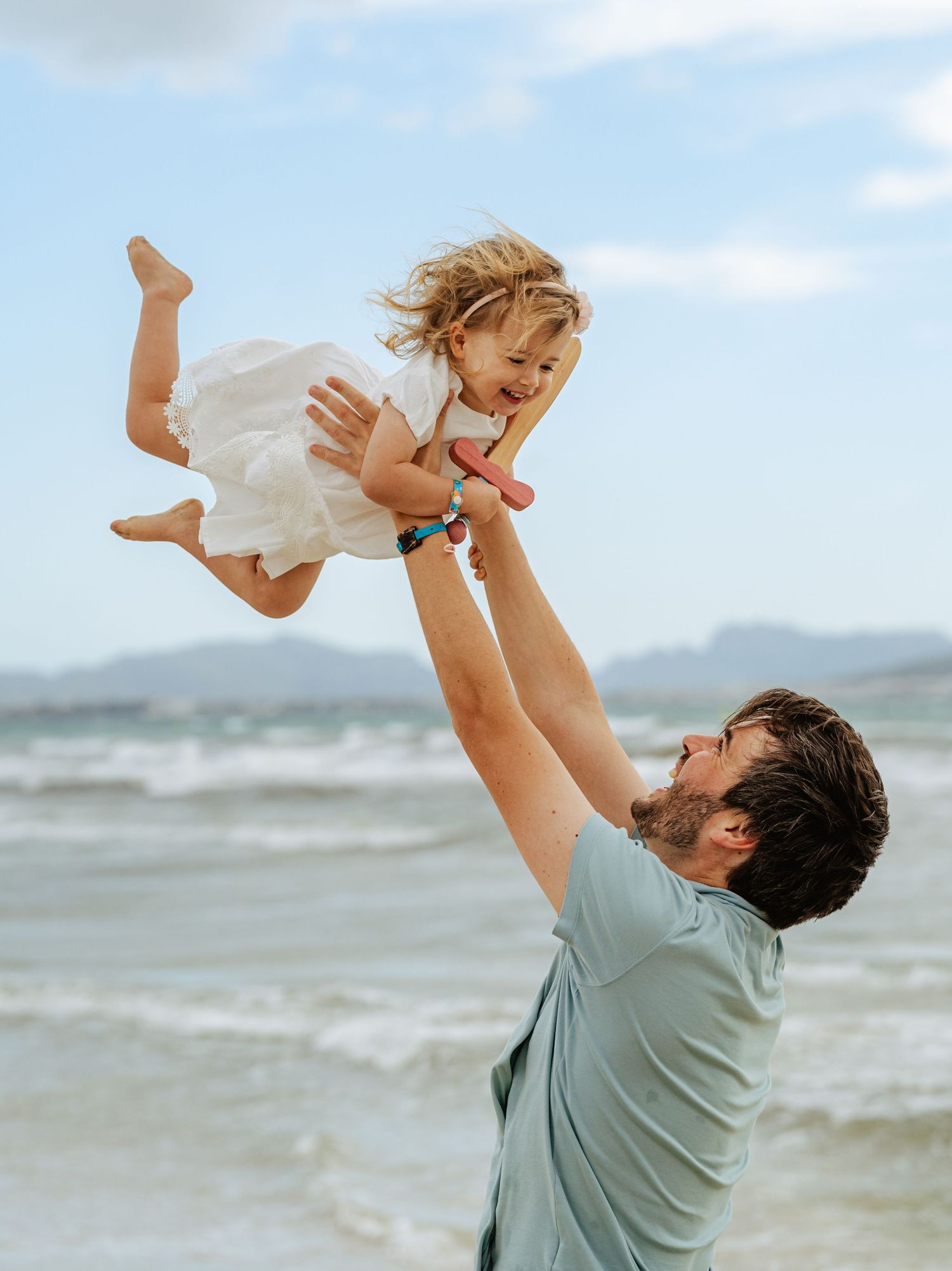 Father joyfully lifting his smiling toddler daughter in the air on a sunny beach.