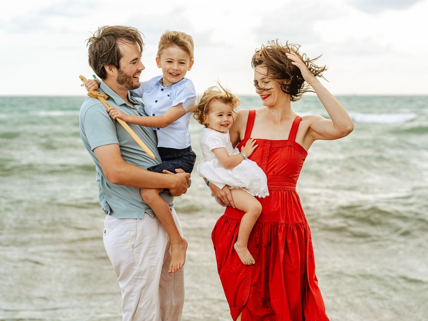 Joyful family of four on a sandy beach, father holding son, mother in red dress holding daughter.