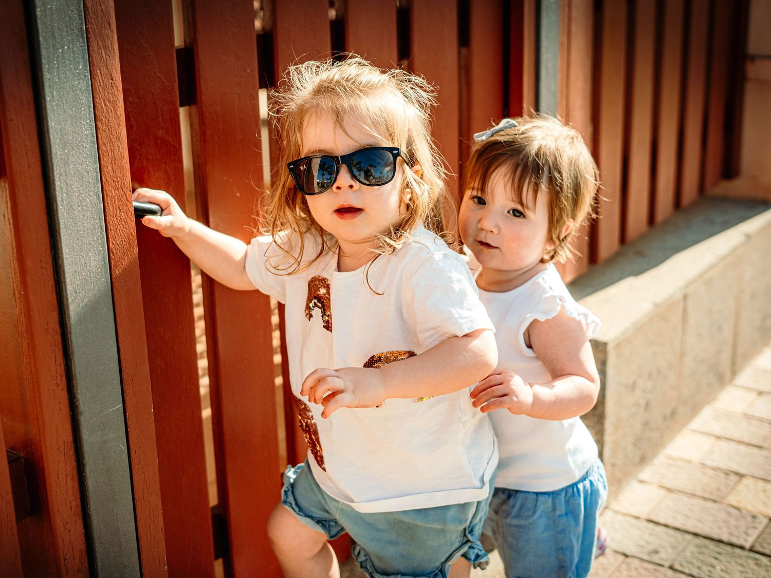 Two young sisters in white tops and denim shorts pose together beside a warm wooden fence outdoors.