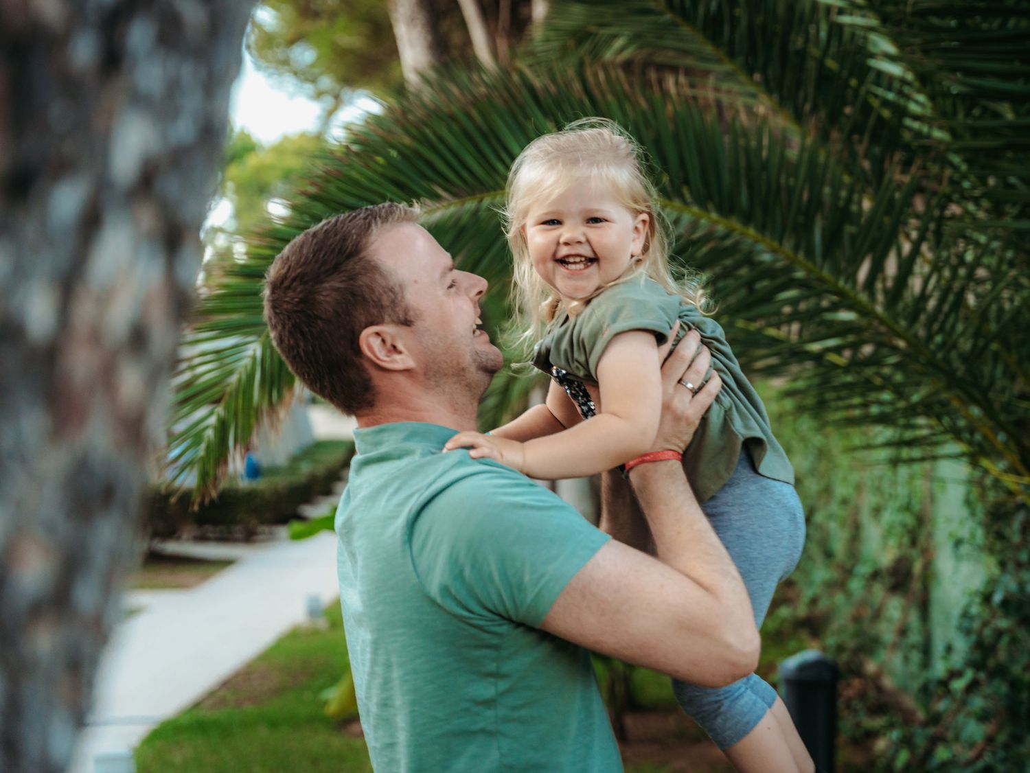 A joyful father in teal shirt lifts his laughing toddler daughter near tropical palm trees.