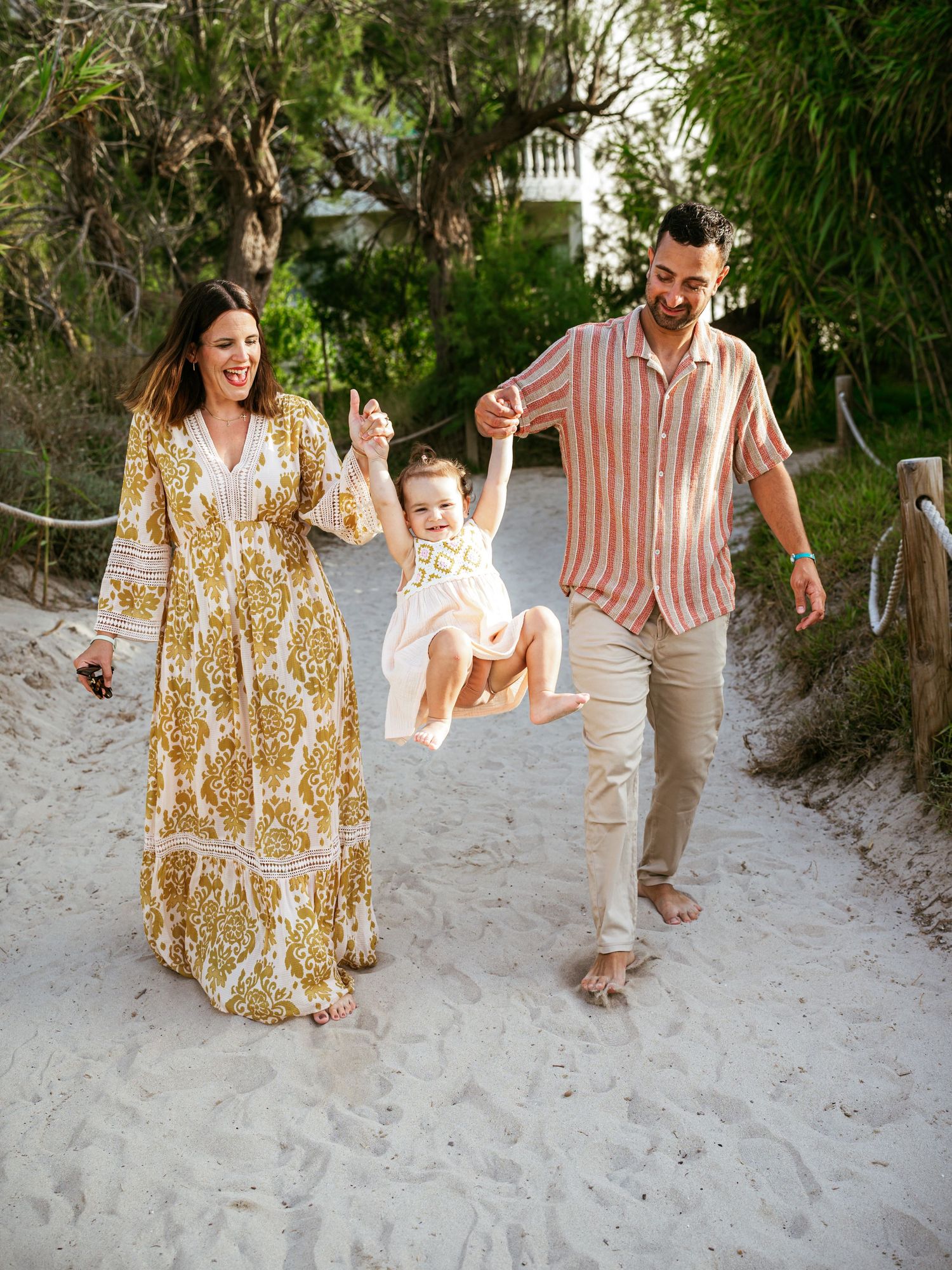 Happy family swinging their baby girl between them while walking on a sandy beach path lined with trees.