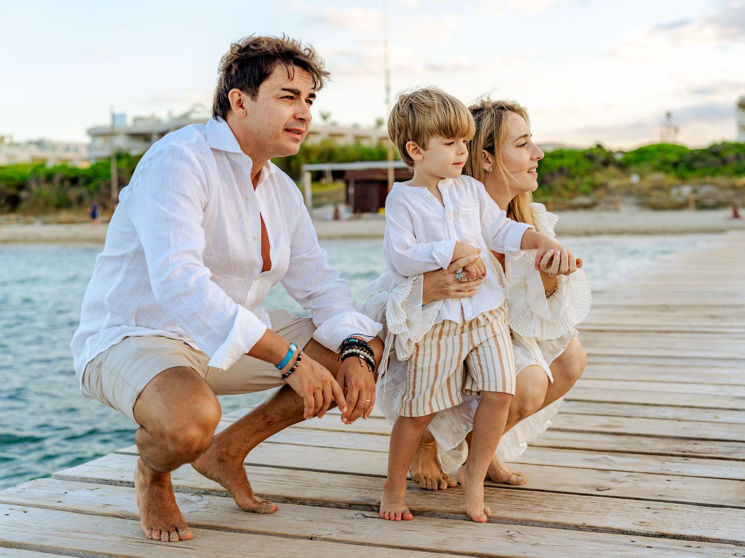 Family of three squatting together on a wooden pier overlooking turquoise ocean at sunset.