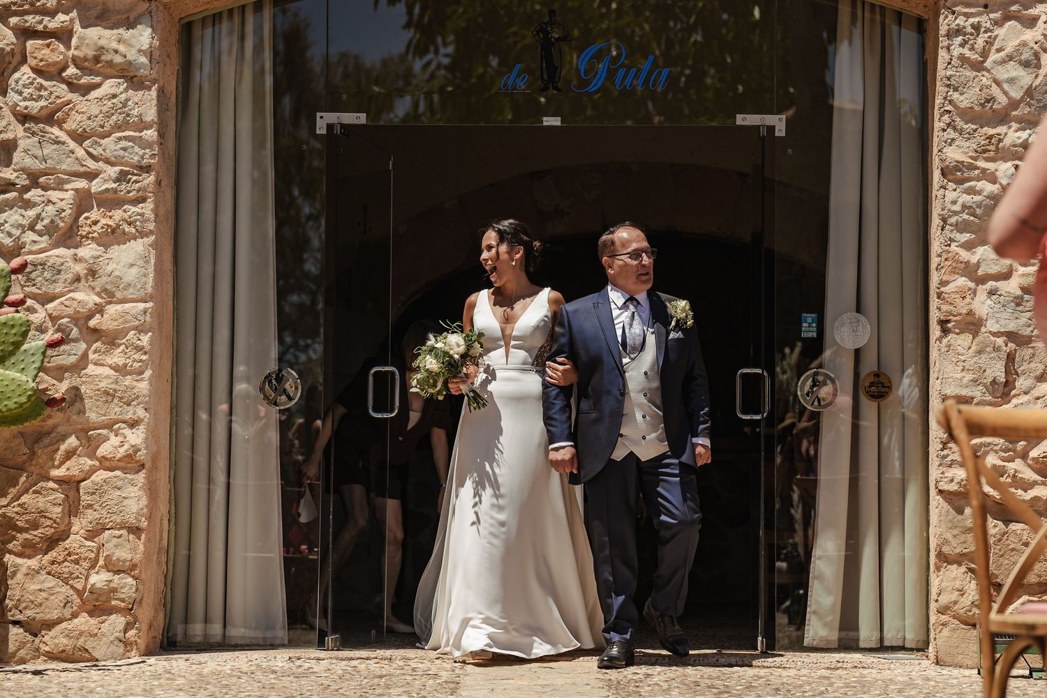Bride in white gown walks with escort through stone building entrance on sunny wedding day.