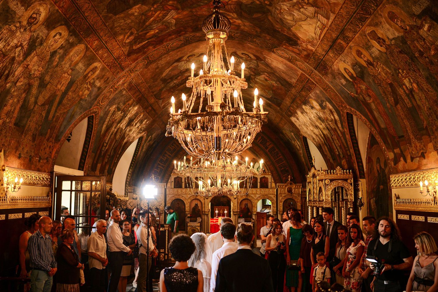 Ornate Orthodox church interior with frescoed ceiling and chandelier filled with wedding guests.