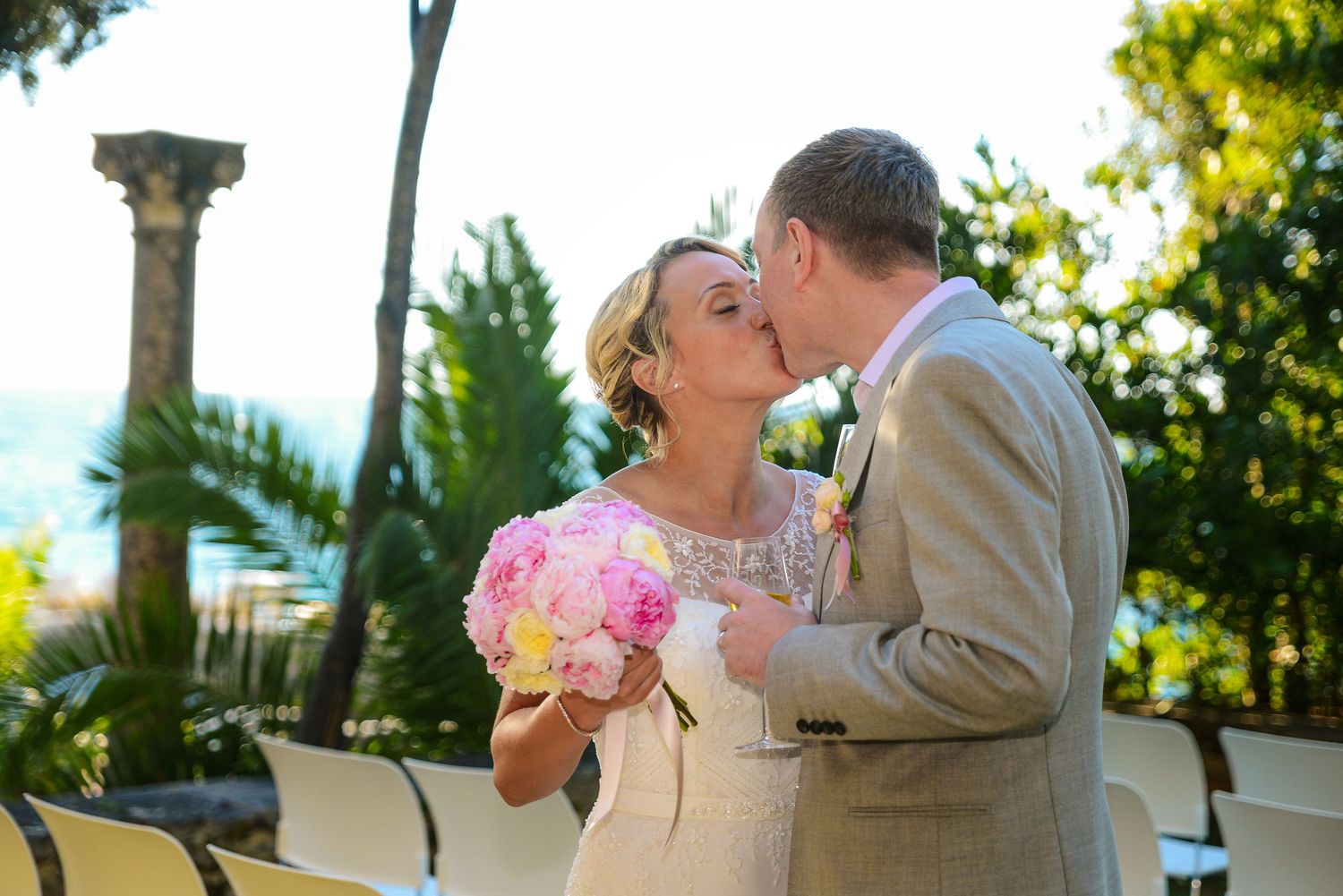 Bride and groom sharing romantic kiss outdoors with pink peony bouquet at Mediterranean wedding.