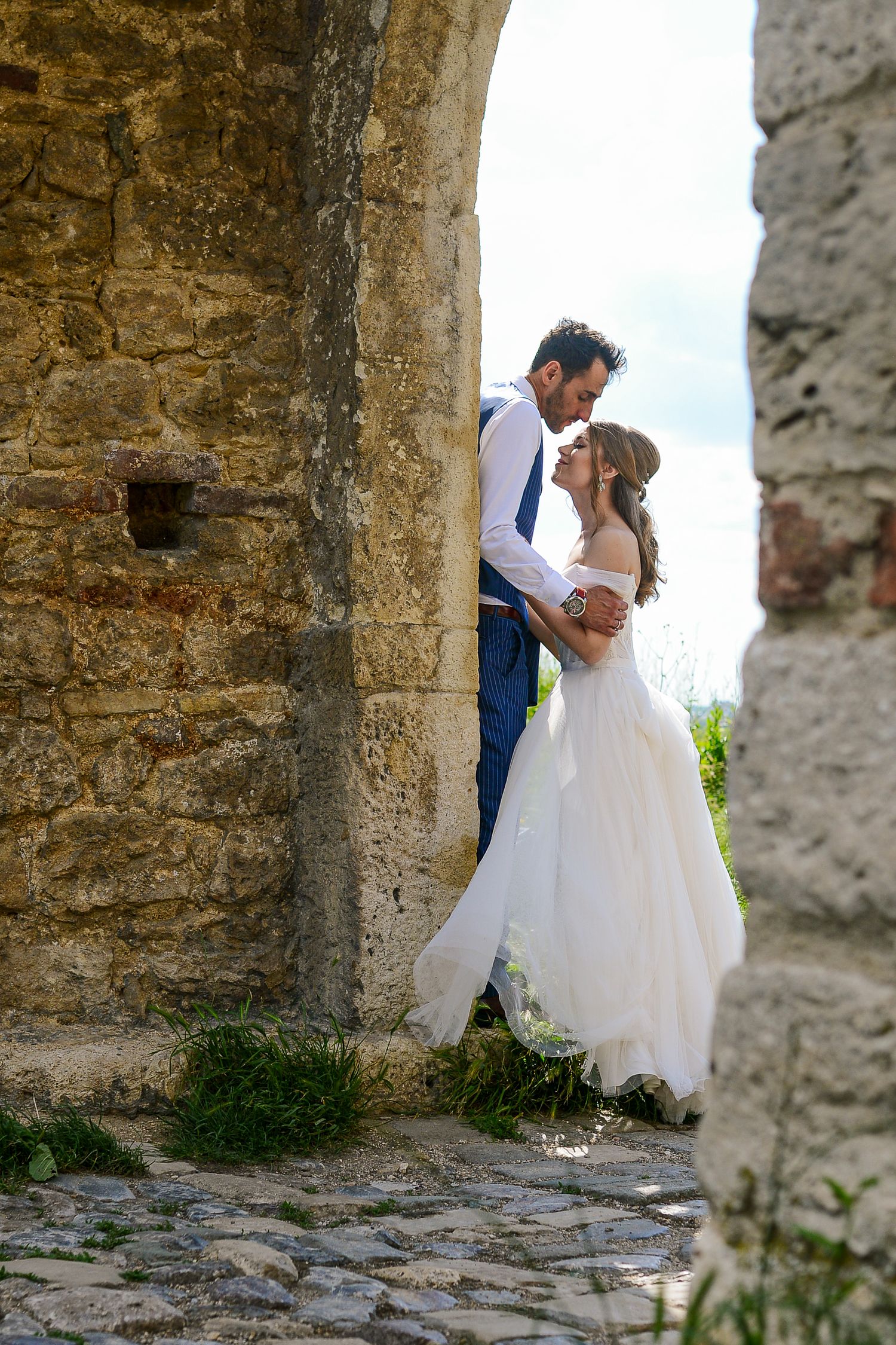 Bride and groom embracing romantically beneath a stone archway at a historic outdoor wedding venue.