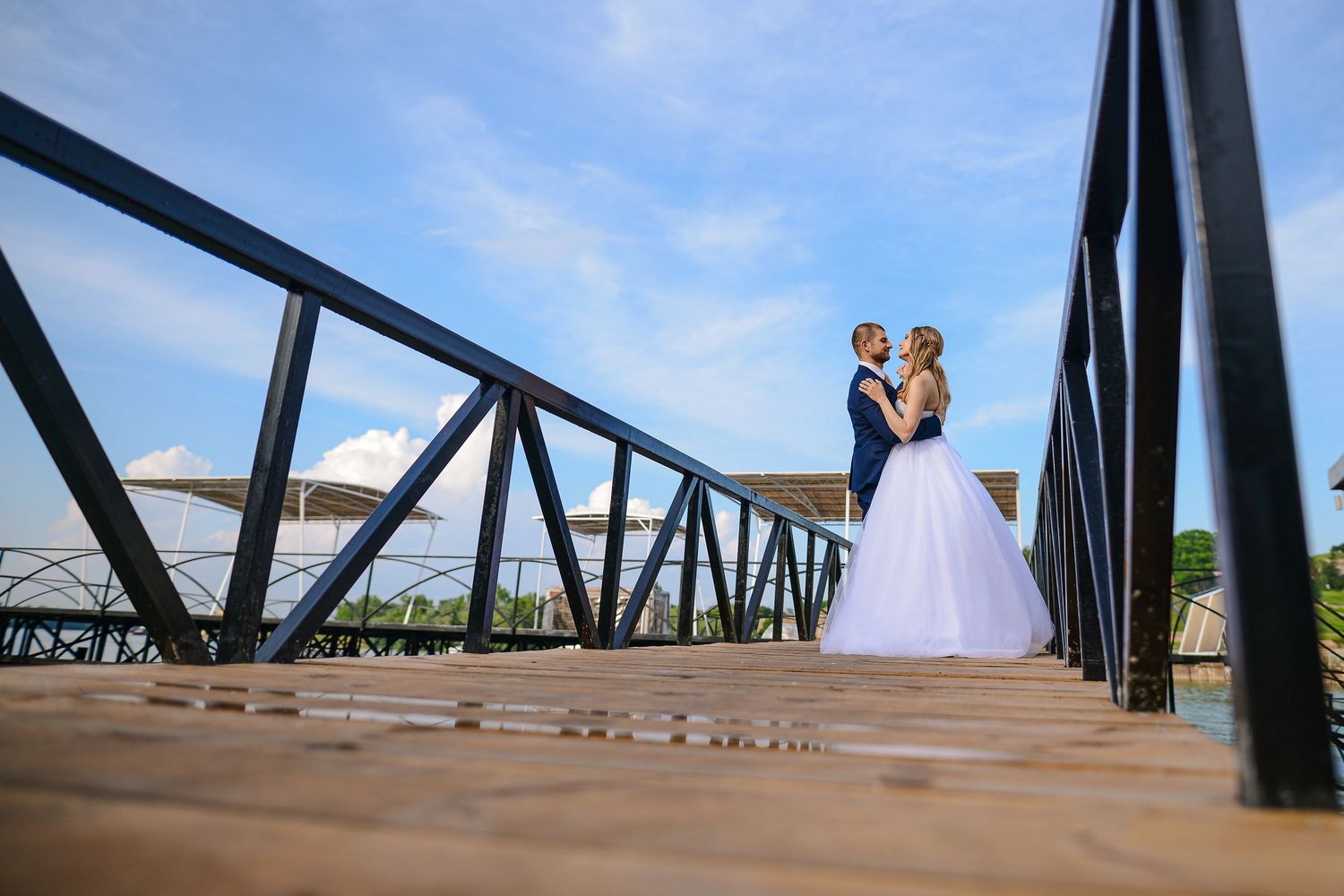 Bride and groom embrace on a wooden bridge under a bright blue sky on their wedding day.