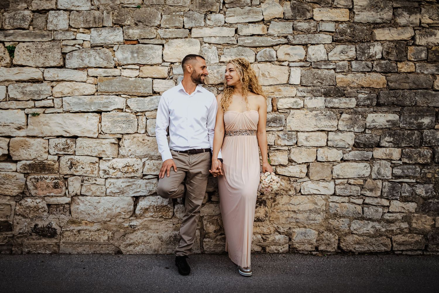 Elegant couple holding hands and smiling at each other against a rustic stone wall backdrop.