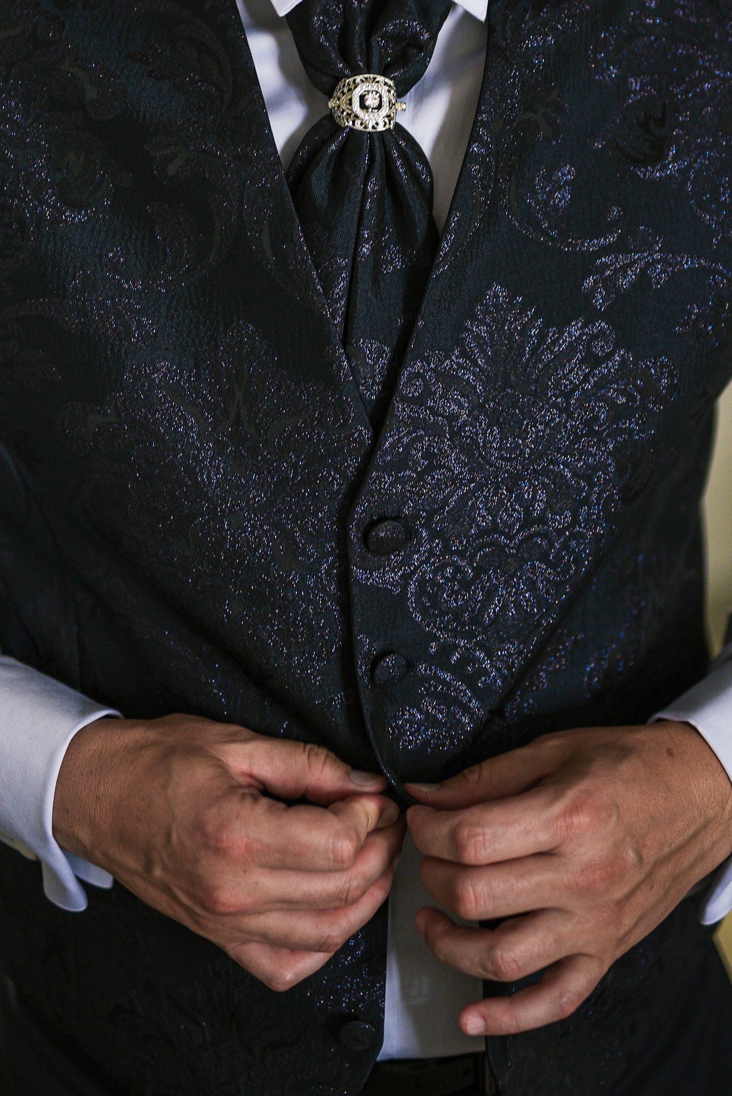 Groom buttoning ornate dark brocade vest with decorative brooch on wedding day.