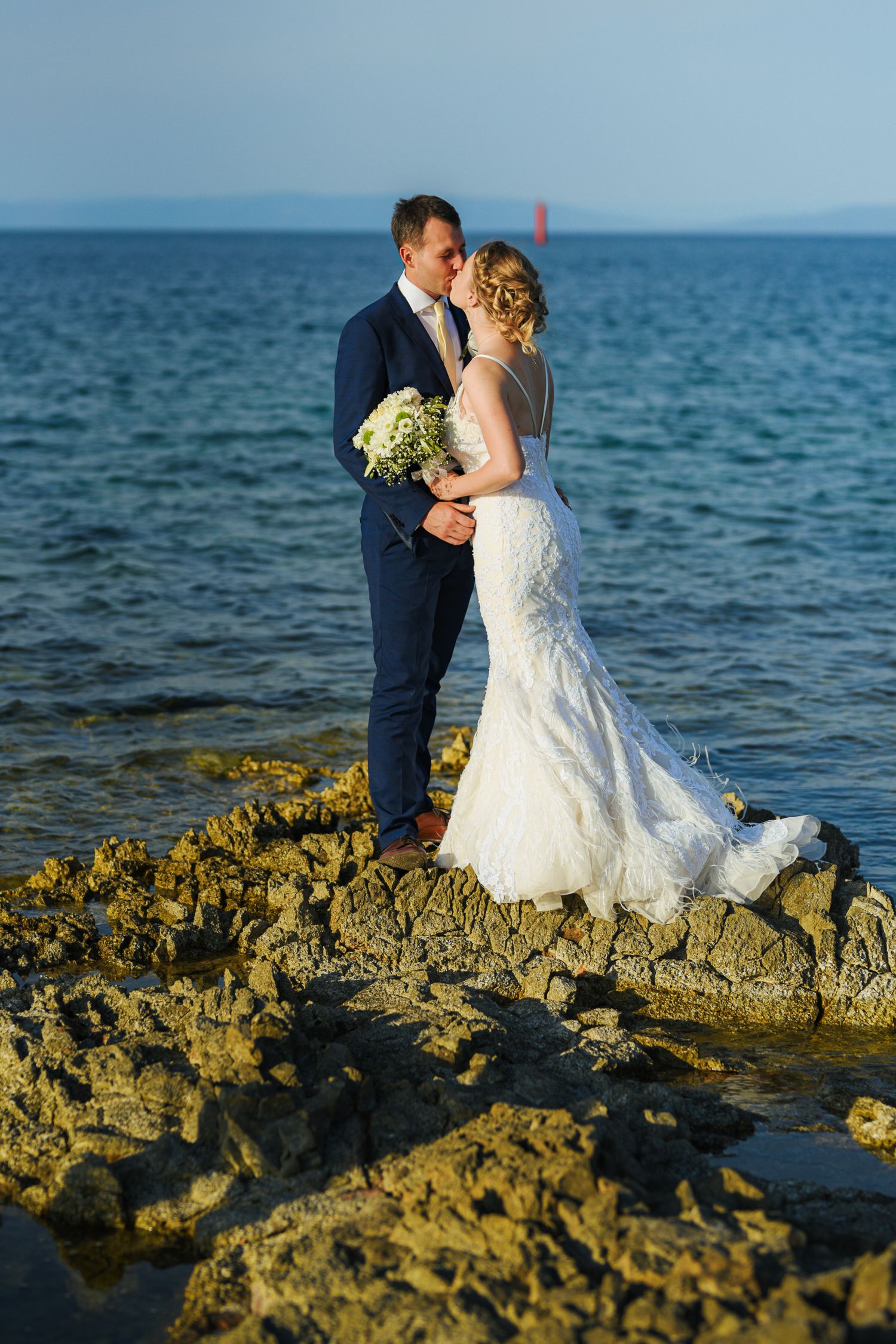 Bride and groom stand romantically on rocky coastal shore, bride holding white bouquet with blue sea behind them.