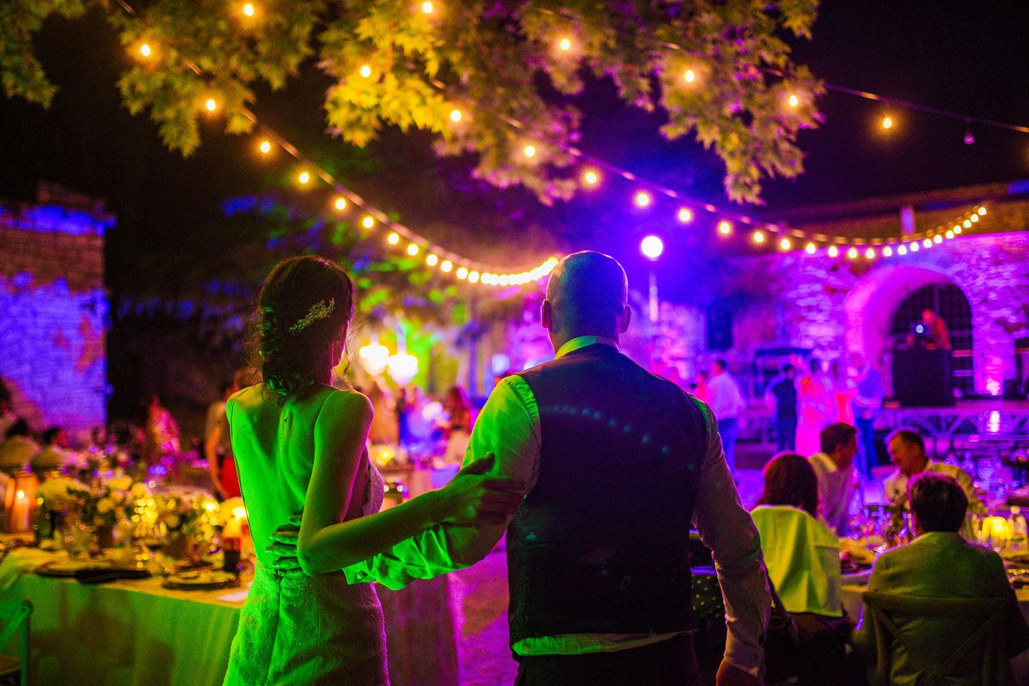 Bride and groom walking together into their colorful outdoor evening wedding reception under glowing string lights. By Herzenshaus Studio.