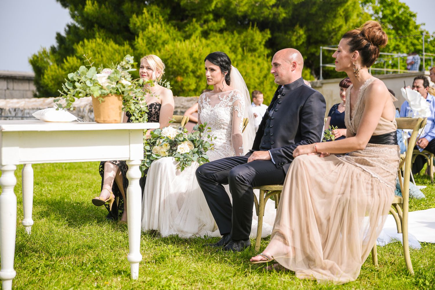 Bride and groom sit with wedding party at outdoor ceremony table on green lawn.