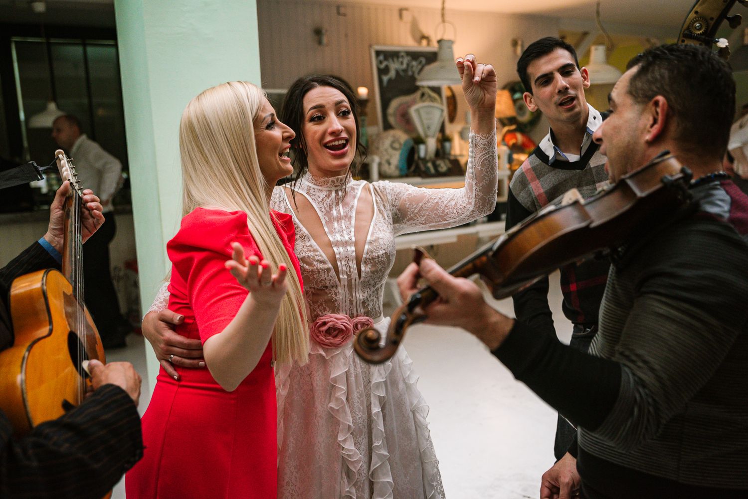 Bride dances happily with blonde guest while musicians play guitar and violin.
