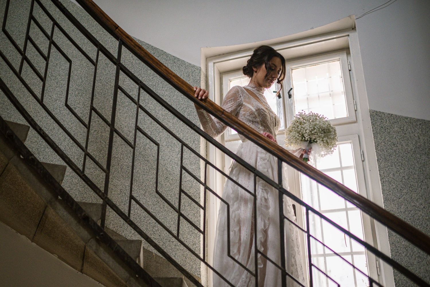 Elegant bride in white lace dress holding small bouquet on staircase landing.