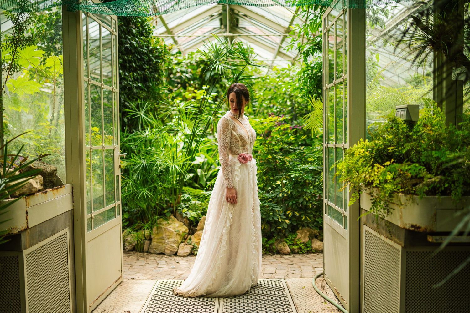 Bride in elegant lace wedding gown standing in doorway of a sunlit greenhouse with tropical plants.