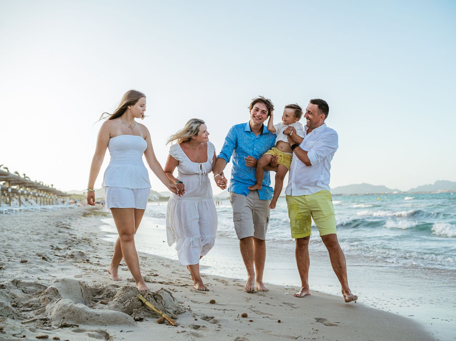 Happy family of five walking together along a sunny beach, laughing and holding hands near the waves.