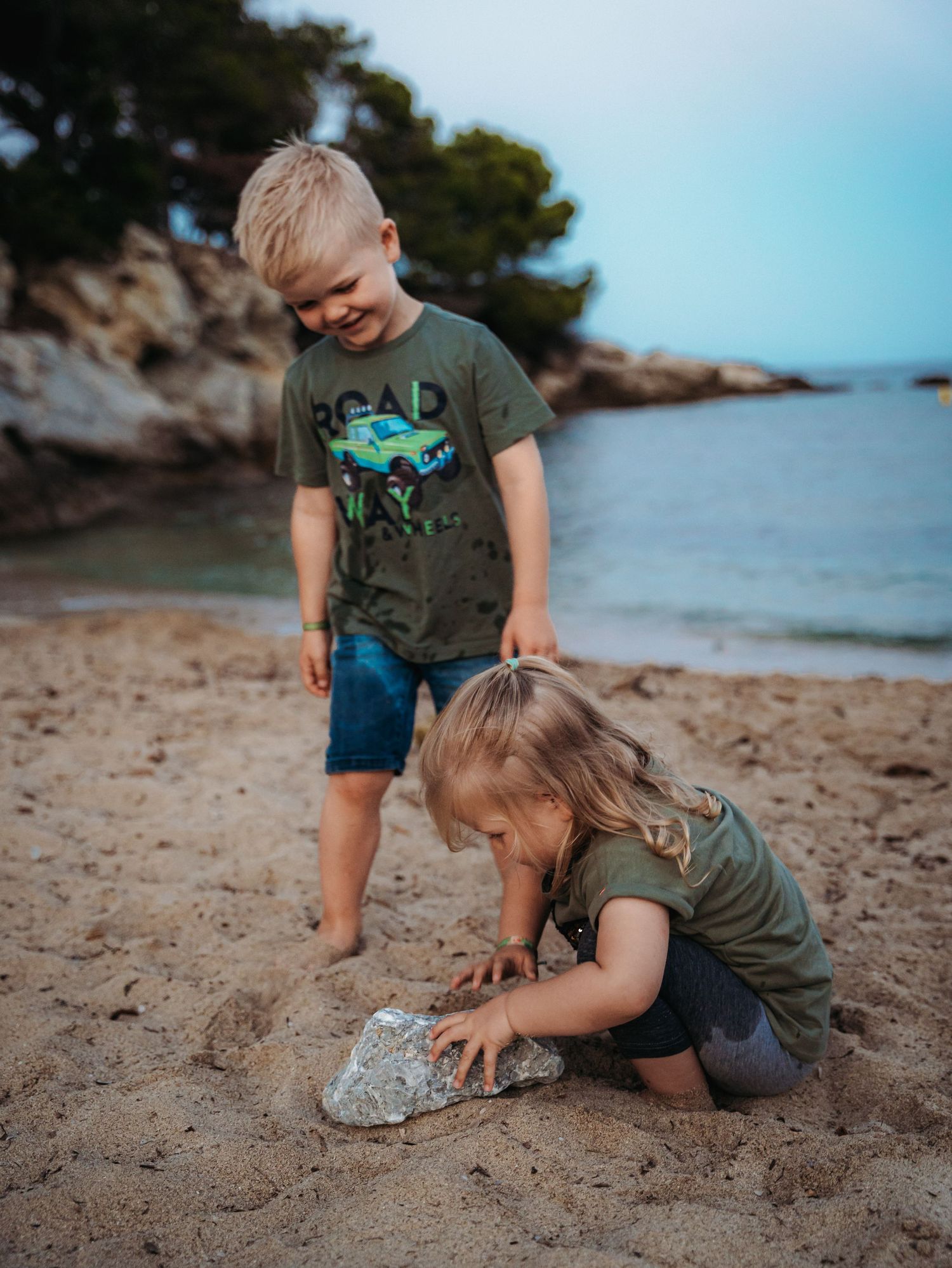 Two young blonde siblings playing together on sandy beach, examining a rock near the water.