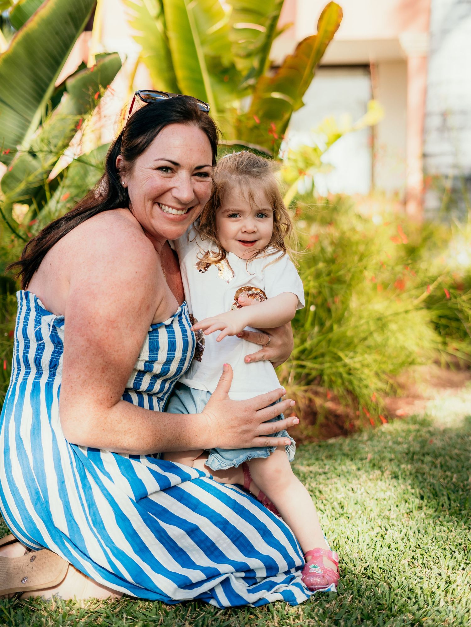 A smiling mother in a blue striped dress sits on grass holding her happy toddler daughter in a tropical garden.