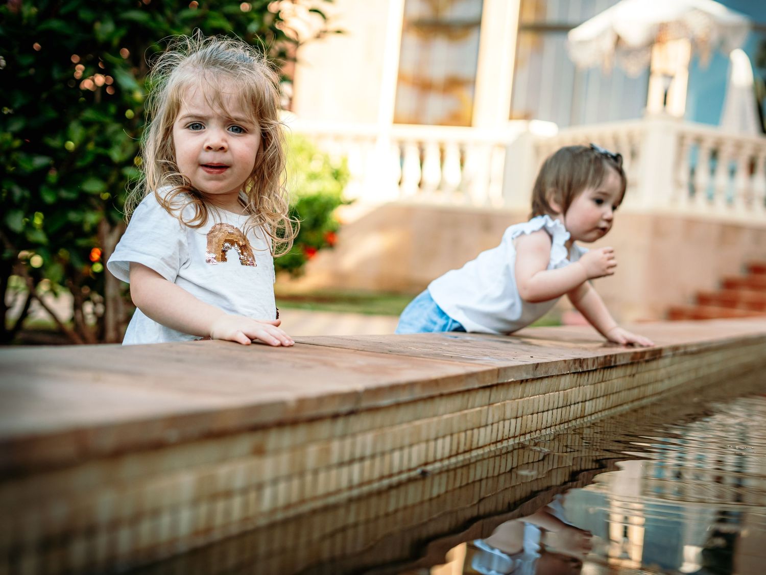 Two young girls in white tops play beside a wooden-edged reflective garden water feature outdoors.