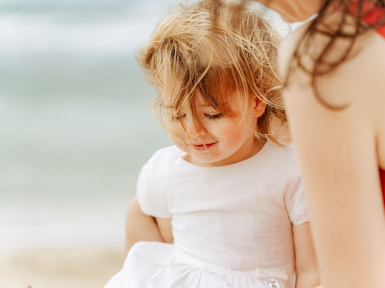 Blonde toddler girl in white dress with windswept hair at the beach near her mother.