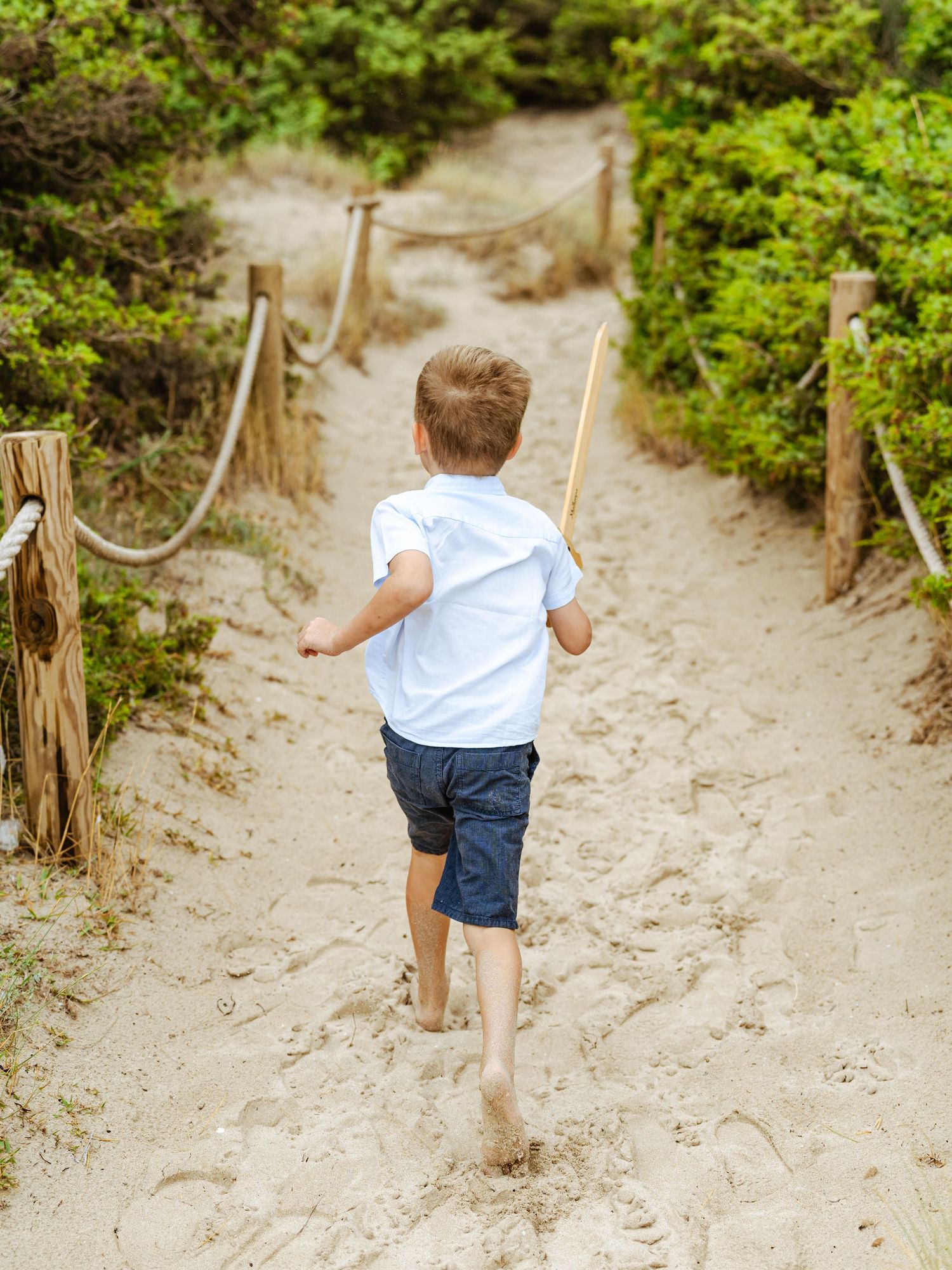 Young boy in white shirt running barefoot down sandy path lined with green bushes.