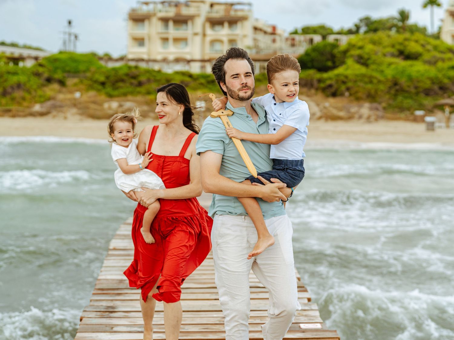 Happy family posing on a seaside wooden dock, parents each holding a young child, waves in background.
