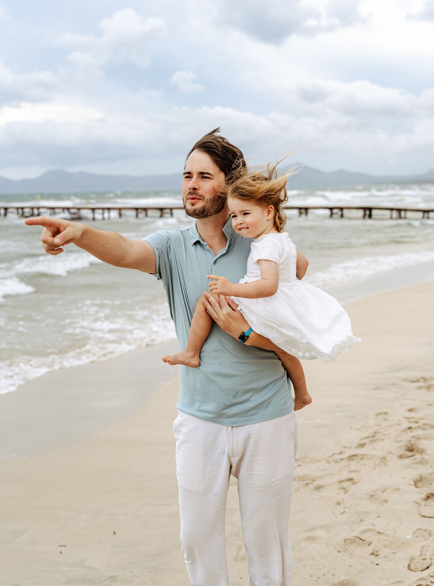 A man in a blue polo holds a little girl in a white dress on a windy beach, pointing into the distance.