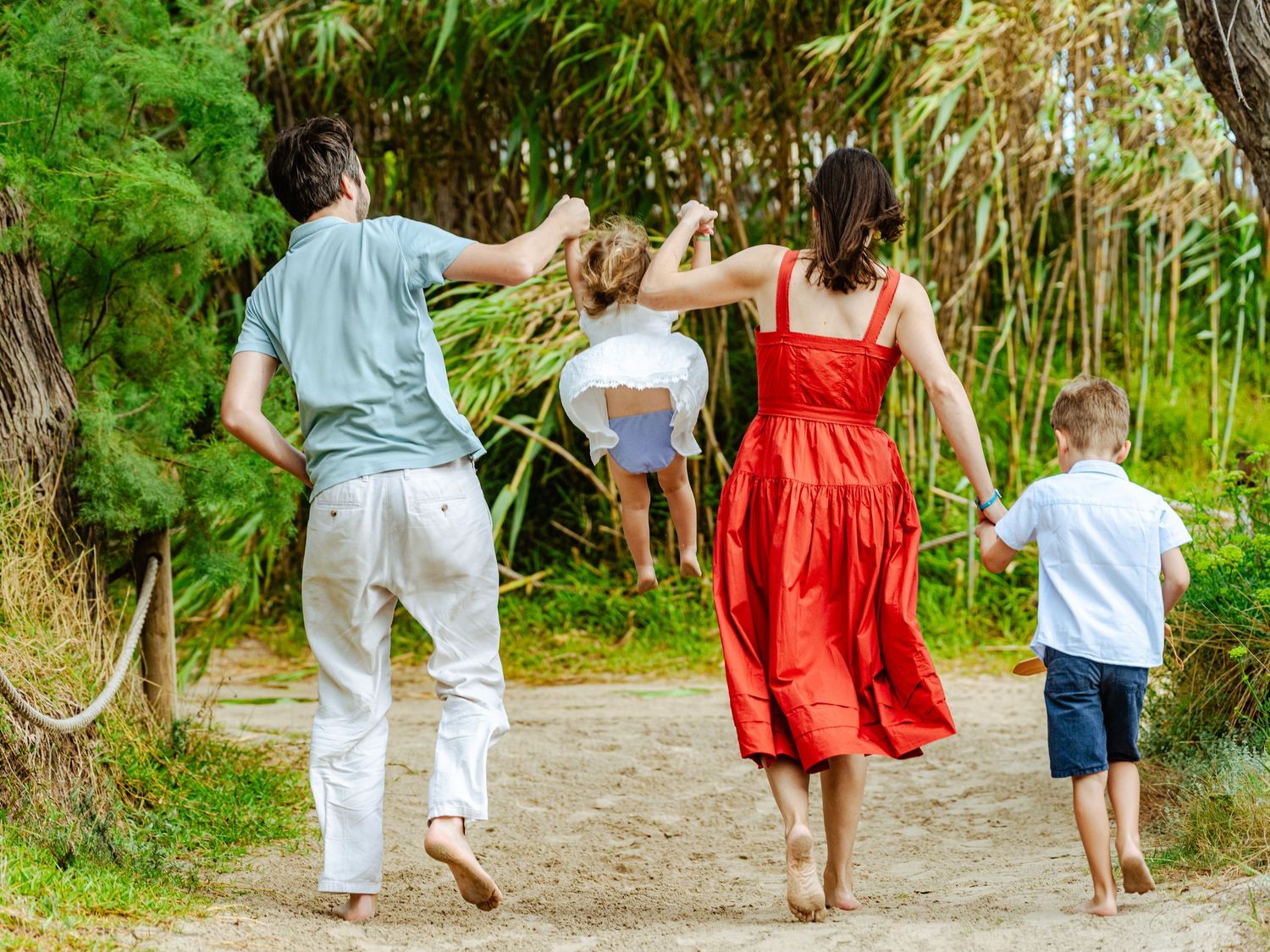 Family of four walking barefoot on sandy trail, parents swinging young girl in the air.