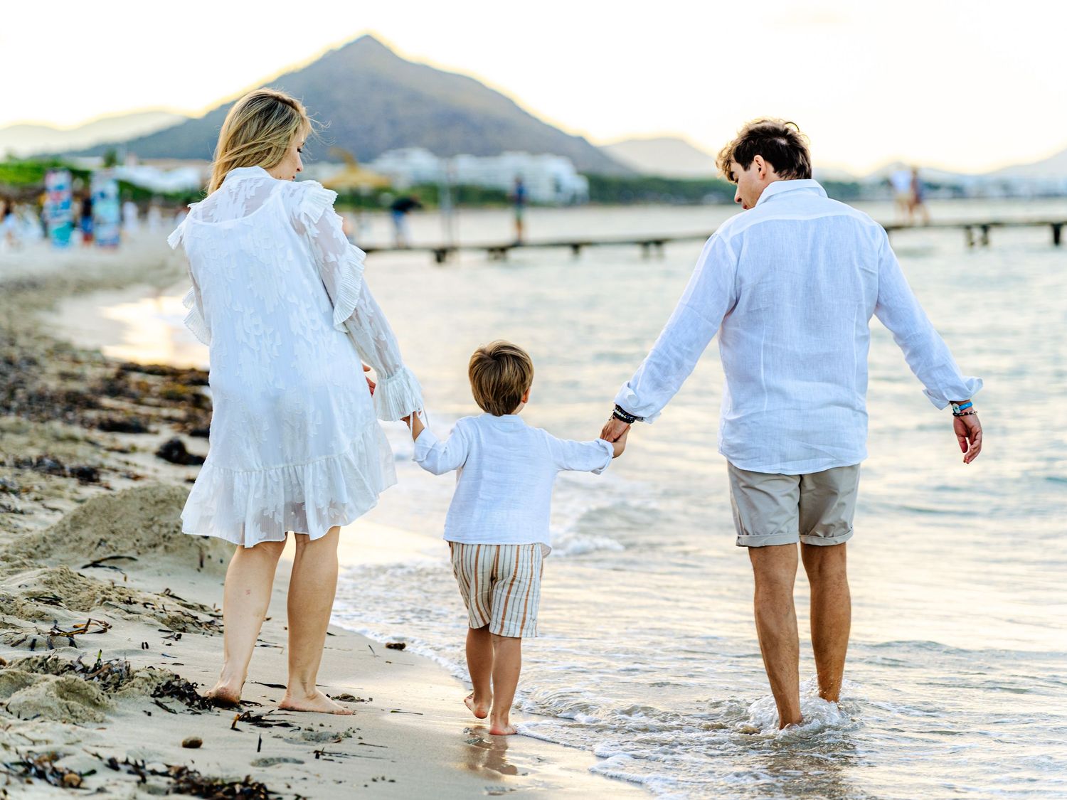 Parents walking hand in hand with their young son along the seashore, captured during a natural family photo session, by Herzenshaus Studio.