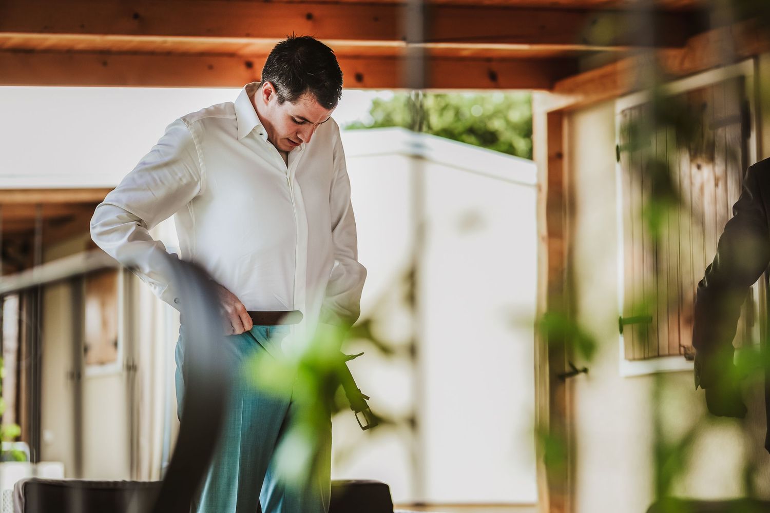 Groom in white shirt adjusting belt in sunlit outdoor venue surrounded by greenery.