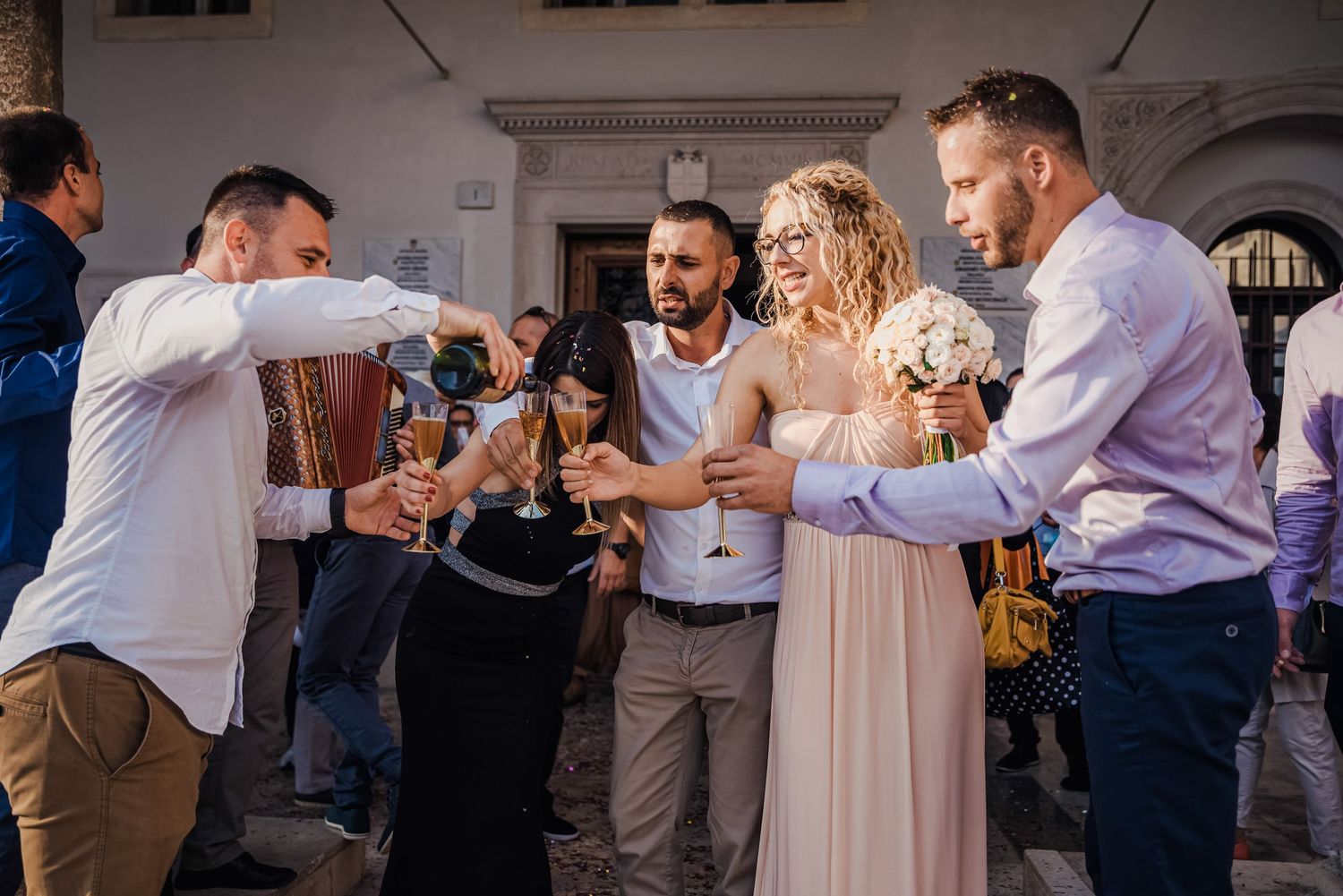 Wedding guests pouring champagne to celebrate with curly-haired bride holding bouquet at outdoor reception.