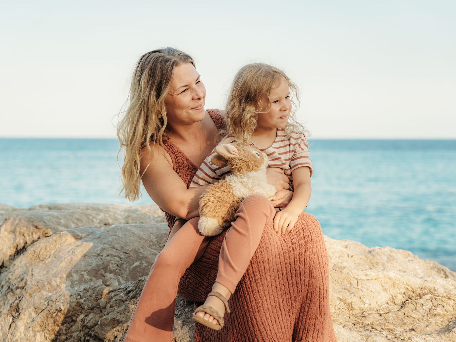 Mother holding young child with stuffed toy on rocky seaside location.