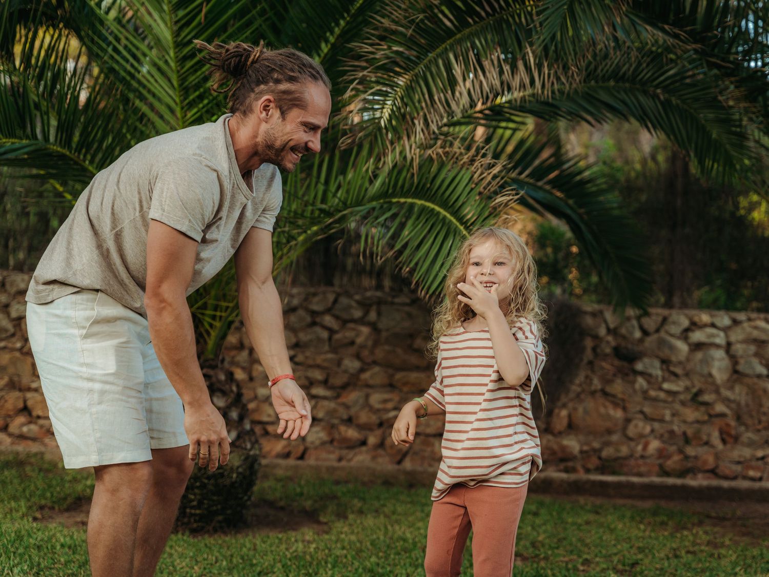 Father playfully interacting with laughing young daughter on grass near stone wall outdoors.