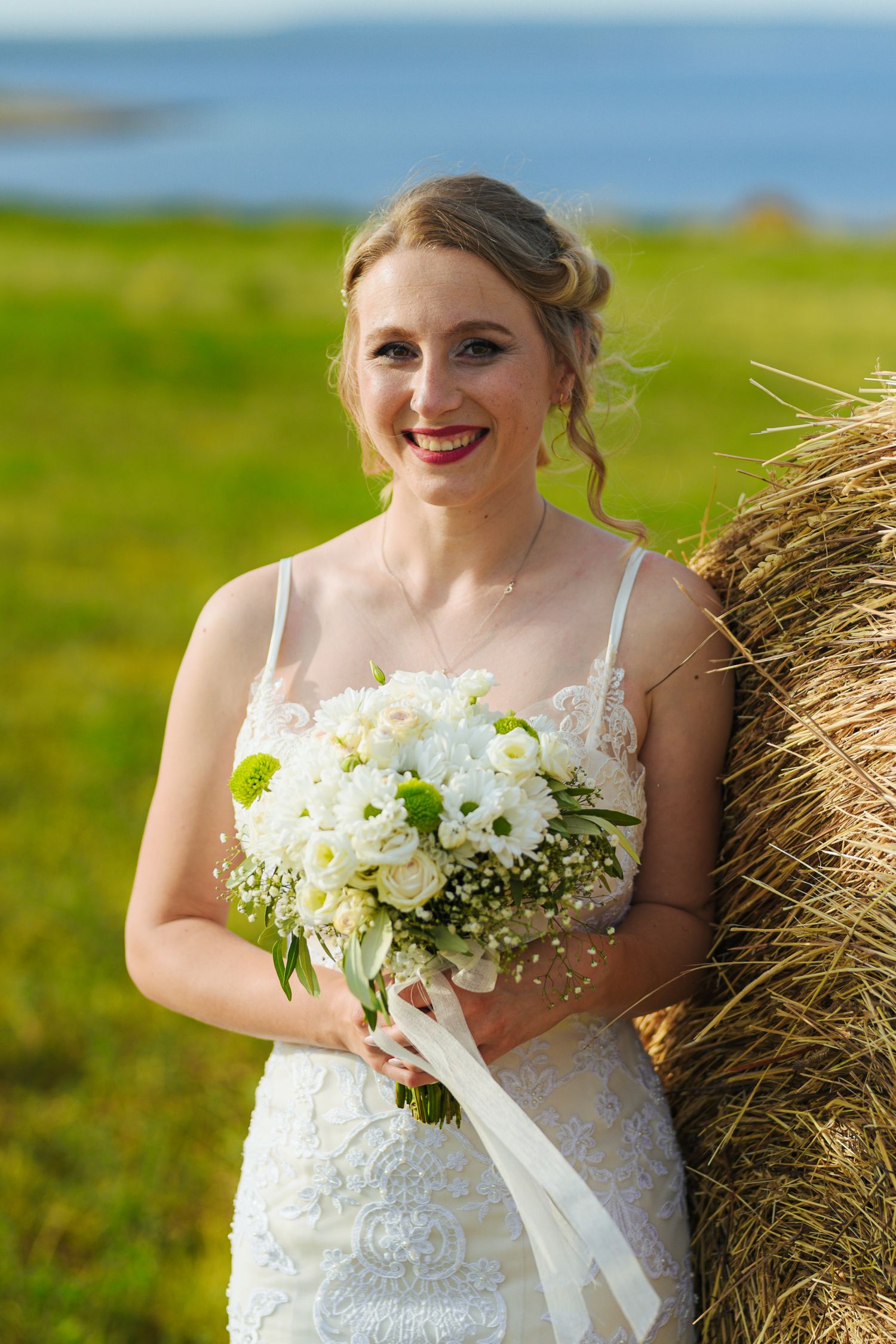 Smiling bride in lace gown holds white bouquet leaning against a hay bale in green field.