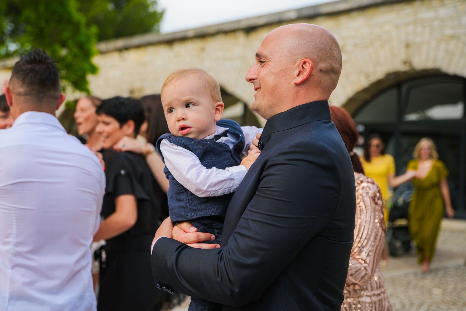 Bald man in navy suit tenderly holds a baby at an outdoor wedding reception.