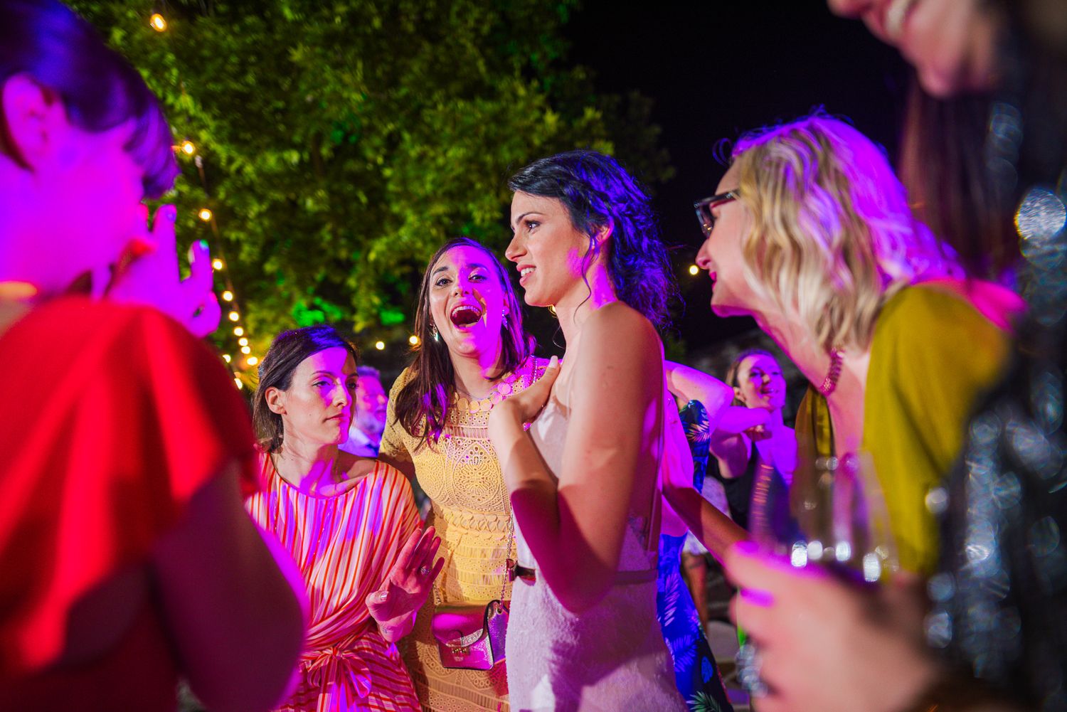 Women laughing and dancing under colorful pink and purple lights at night wedding.