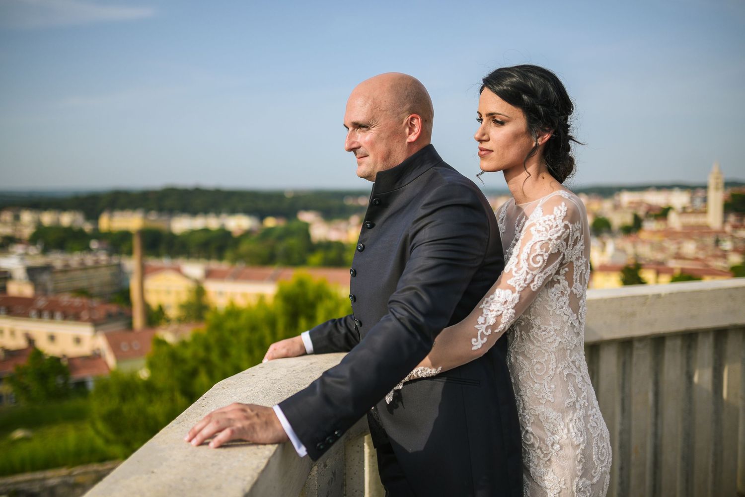 Bride and groom standing on a balcony overlooking a European city at golden hour.