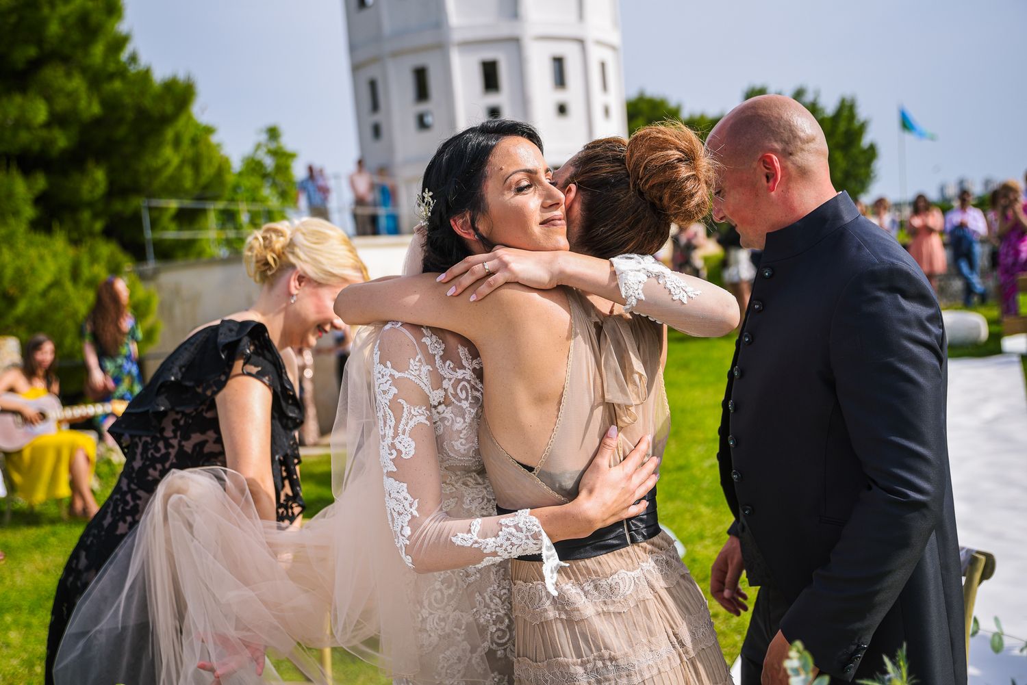 Bride in white lace gown sharing warm hugs with guests near a white tower at outdoor wedding.