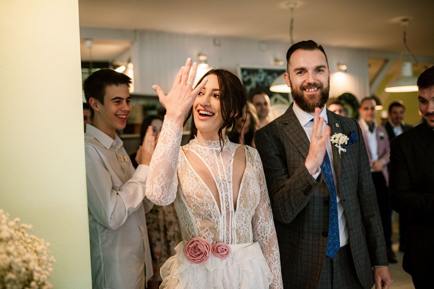 Radiant bride in lace gown waves to guests alongside smiling groom at wedding reception.