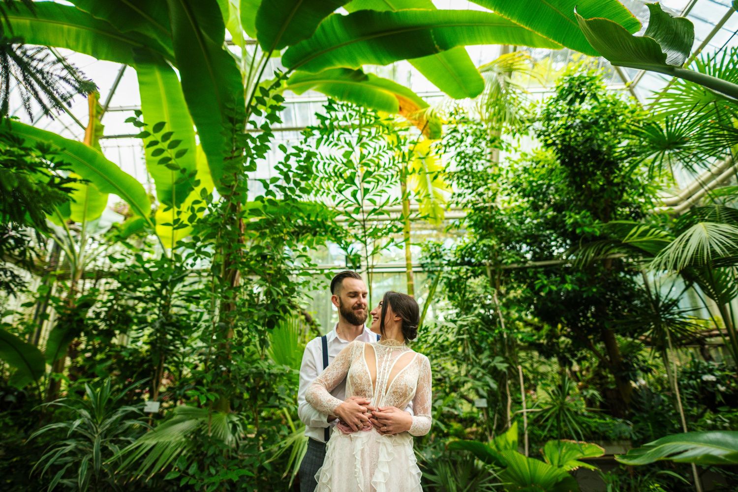 Bride and groom stand together surrounded by lush tropical plants inside a botanical greenhouse.