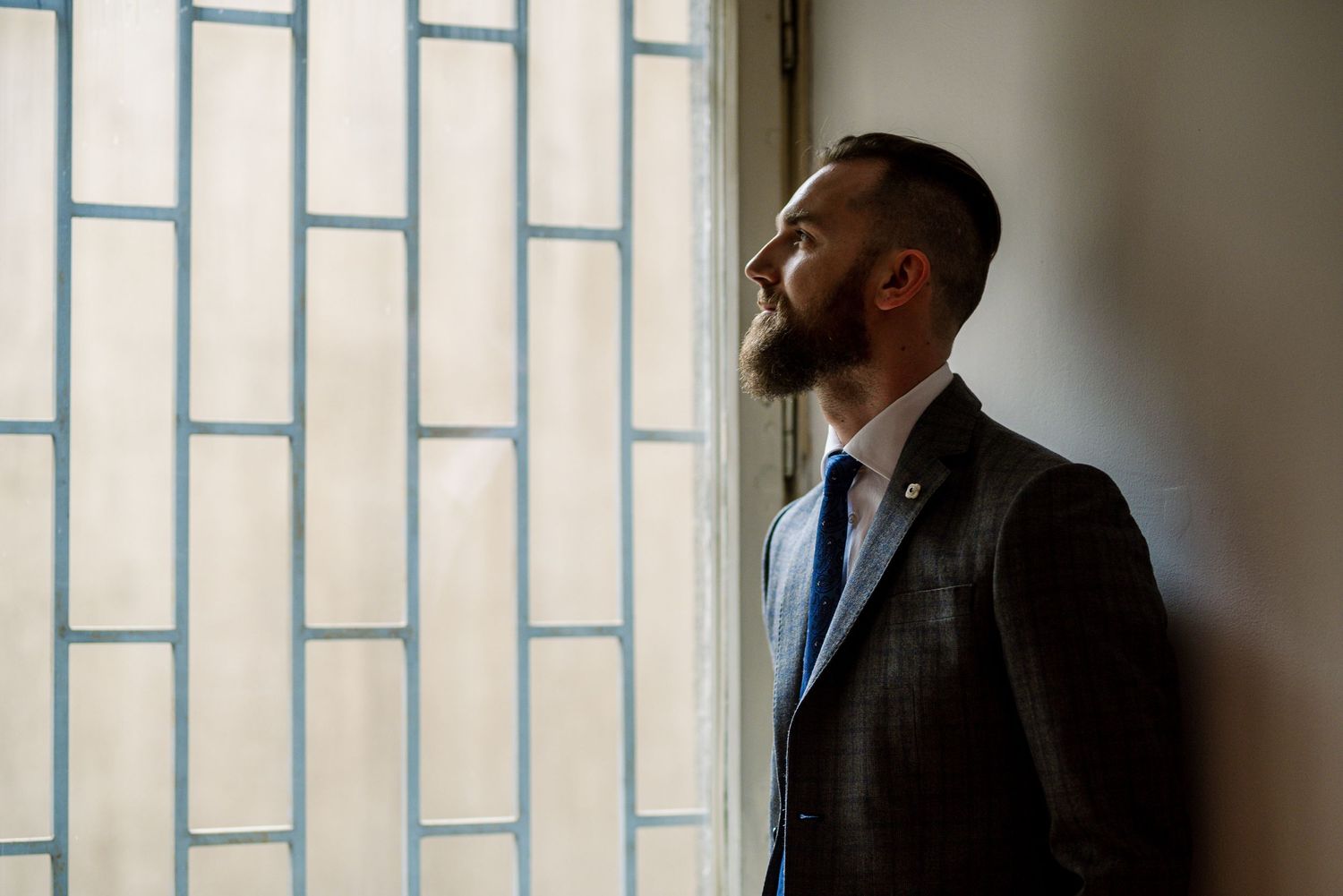 Bearded groom in suit gazing upward by stained glass window on wedding day.